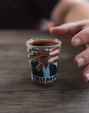 Trump Shot Glass - Trump Shot Glass.  Casual Trump pictured in a red MAGA hat, pointing, American flag design in the background.  One ounce.